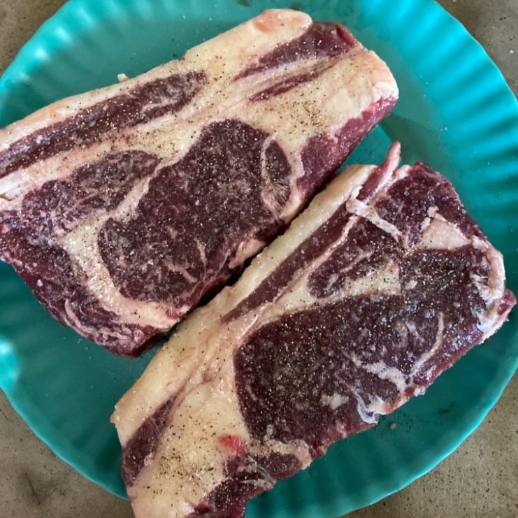 Two seasoned ribeye steaks displayed on a turquoise plate, showcasing marbled red meat and white fat, ready for cooking.