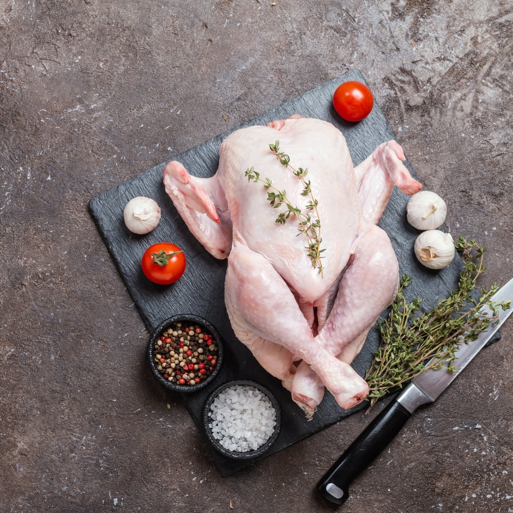 Raw whole chicken on a slate cutting board, garnished with fresh thyme, surrounded by cherry tomatoes, garlic, and bowls of mixed peppercorns and sea salt, with a chef's knife nearby.